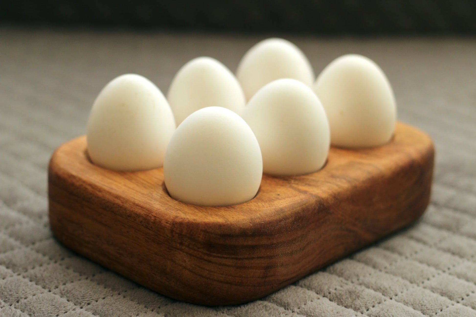 Top view of solid wood egg tray with six round slots holding brown eggs on a kitchen counter.