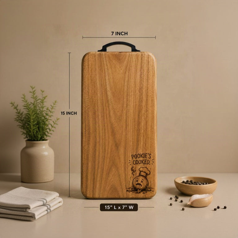 A wooden cutting board with a black handle, shown with dimension lines indicating 15 inches by 7 inches. The board has a warm wood grain finish and a black engraving that reads 'POOKIE'S COOKED.