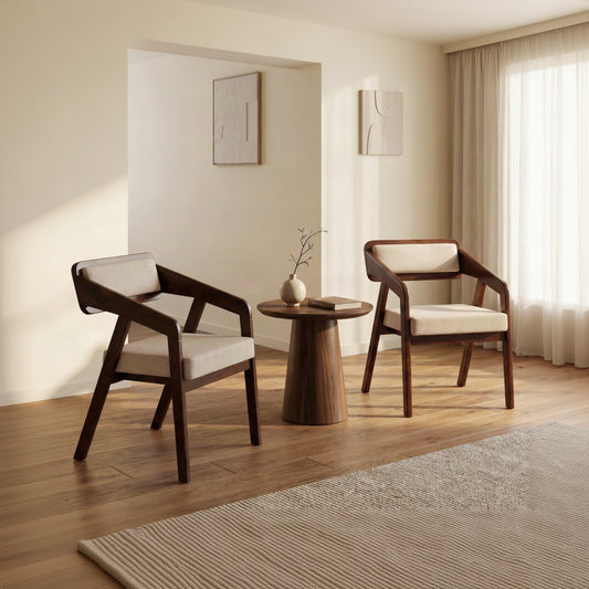 Two modern wooden armchairs with cream cushions and a small round wooden side table in a bright, minimalist room with warm sunlight and curtains.