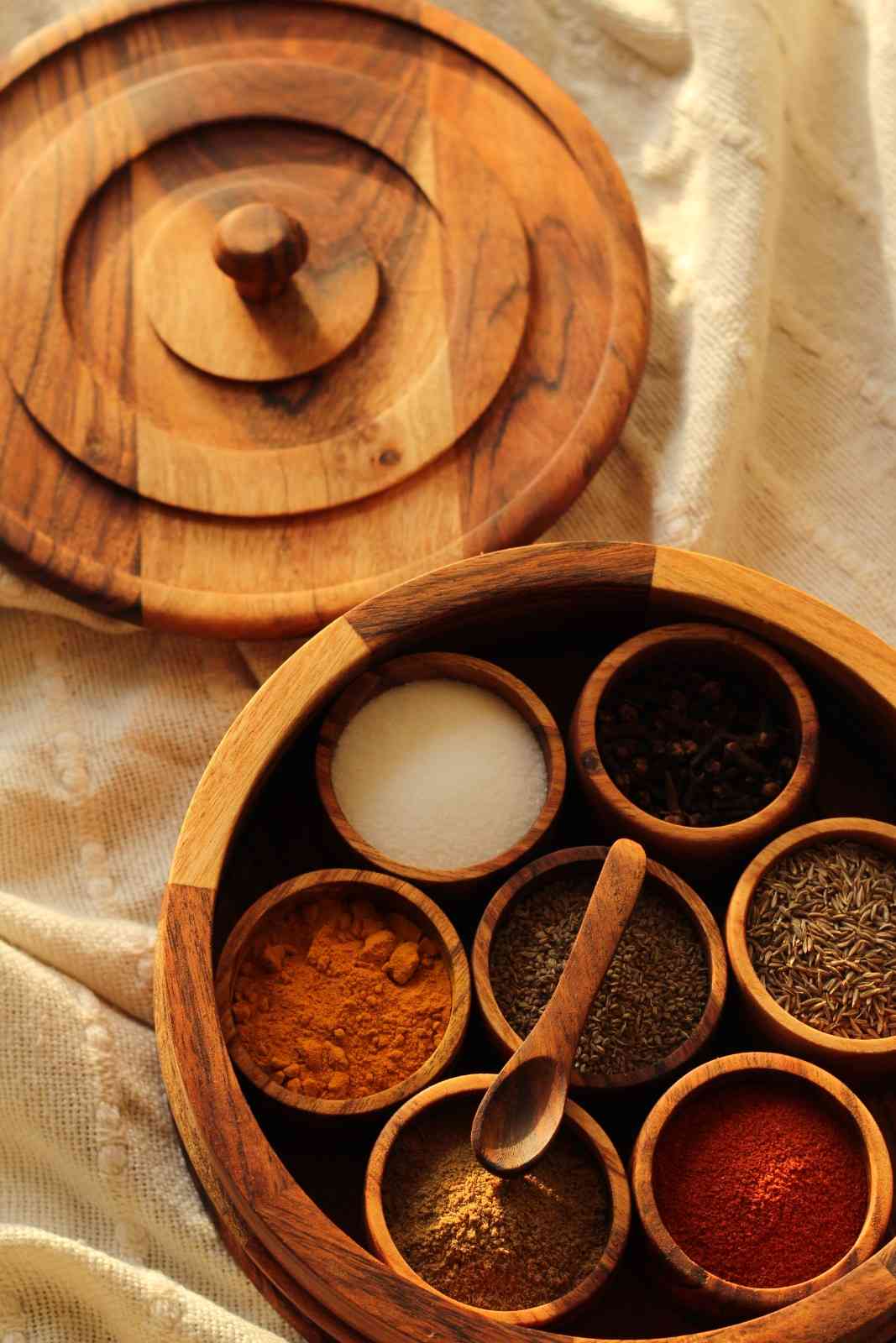 Wooden spice rack with various spices and a wooden spoon on a beige fabric background